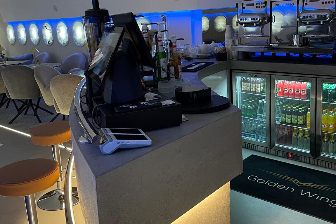 The finished bar: grey stone-topped counter with the Golden Wings name inset in the front face, espresso machine and grinders on the back bar, glass-front drinks fridge, leather-topped stools, portholes and blue cove behind.