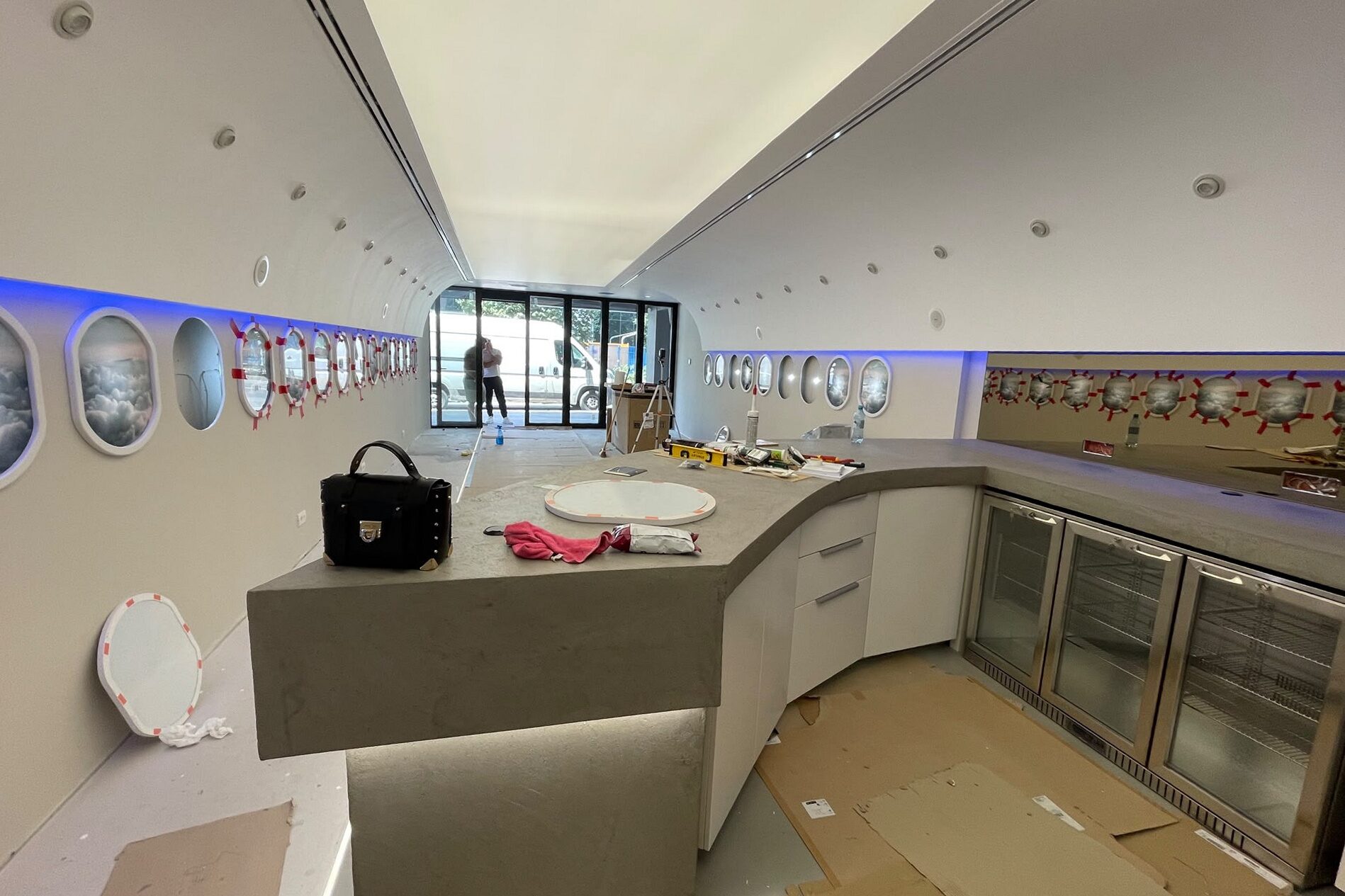 The bar under construction: curved grey counter run built out with glass-front under-counter fridges installed, glazed portholes and lit blue cove along both walls, floor still protected with board.