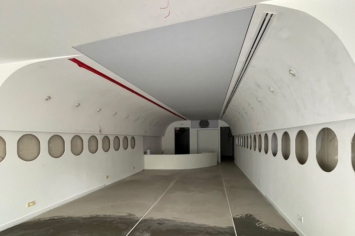 The bare fuselage shell looking down the length of the space: curved plasterboard ceiling arc springing from both side walls, porthole openings unglazed, red accent stripe on the crown, raw concrete floor.