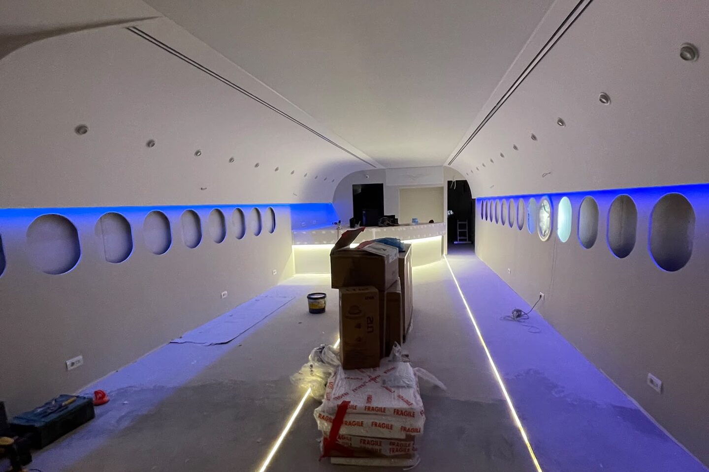 Cove and floor lighting under test before fit-out: blue LED cove washing the porthole band on both walls, two warm LED strips running the length of the floor, a wrapped pallet of materials mid-span.