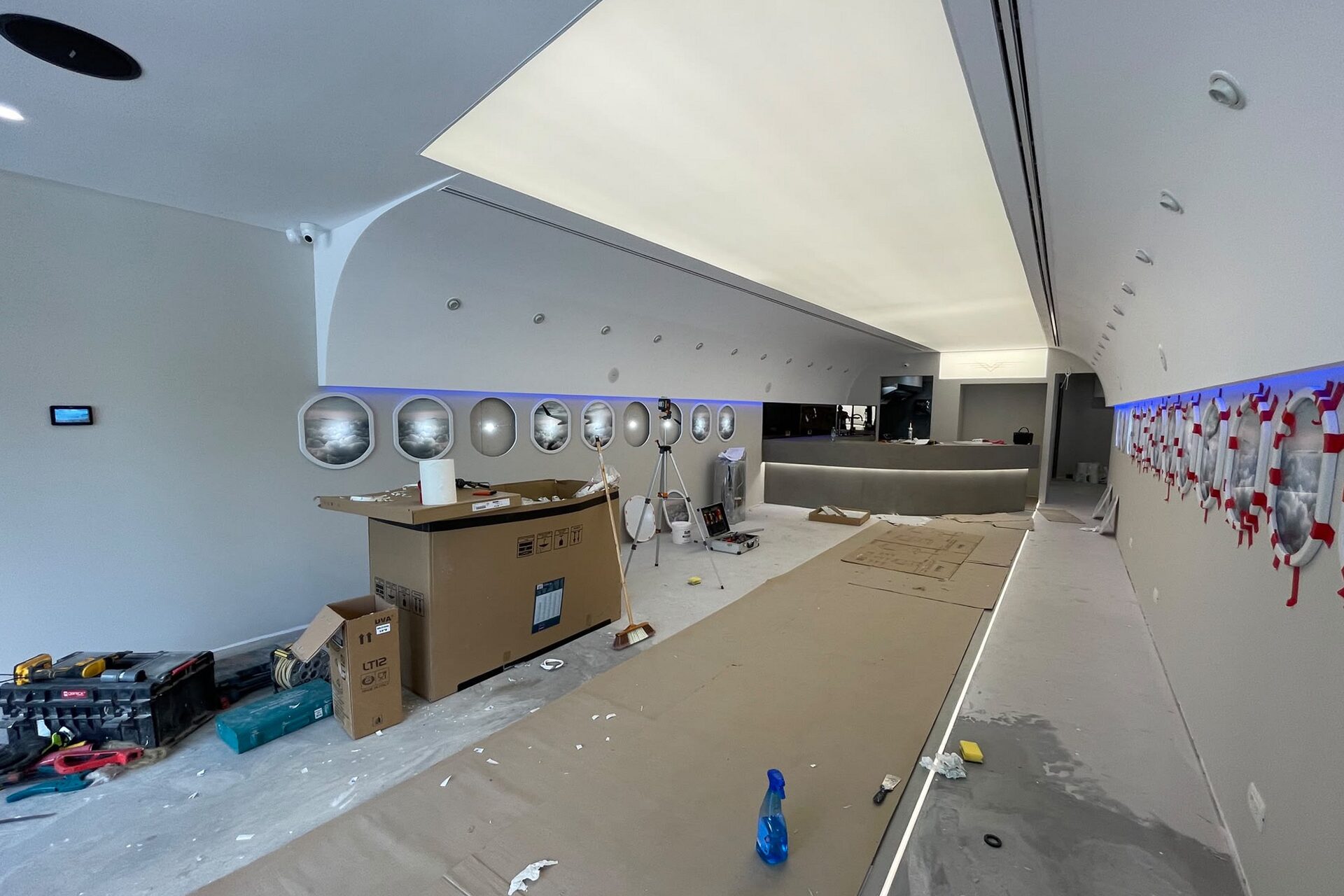 Porthole glazing being set: red spring clamps holding freshly bonded oval surrounds along the right wall while the adhesive cures, service counter carcass built out on the left, ceiling arc complete.