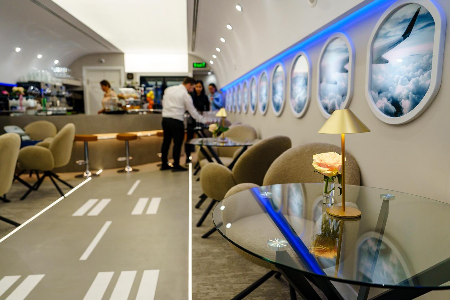 Cabin seating in use: backlit portholes showing cloud and wingtip imagery above a run of boucle chairs and glass tables, brass table lamp in the foreground, runway markings underfoot, bar and staff beyond.