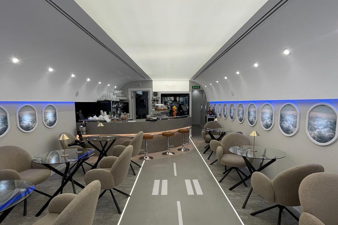 The finished room down its centreline: runway threshold markings and centreline painted on the floor, glazed portholes with backlit cloud graphics and blue cove down both walls, upholstered chairs at glass tables, bar at the far end.