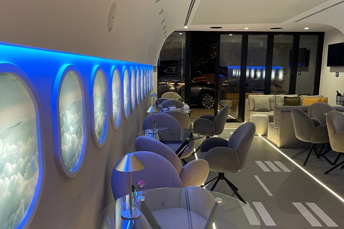 Looking along the porthole wall at night: blue cove light over backlit cloud portholes, glass-topped tables on black star bases, boucle chairs, runway markings on the floor and the glazed end wall beyond.