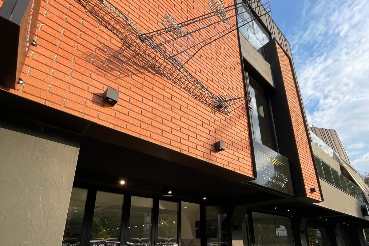 Street elevation with the outdoor terrace in use: wire aeroplane sculpture on the brick facade above, cafe tables and chairs on the paved terrace below the glazed frontage.