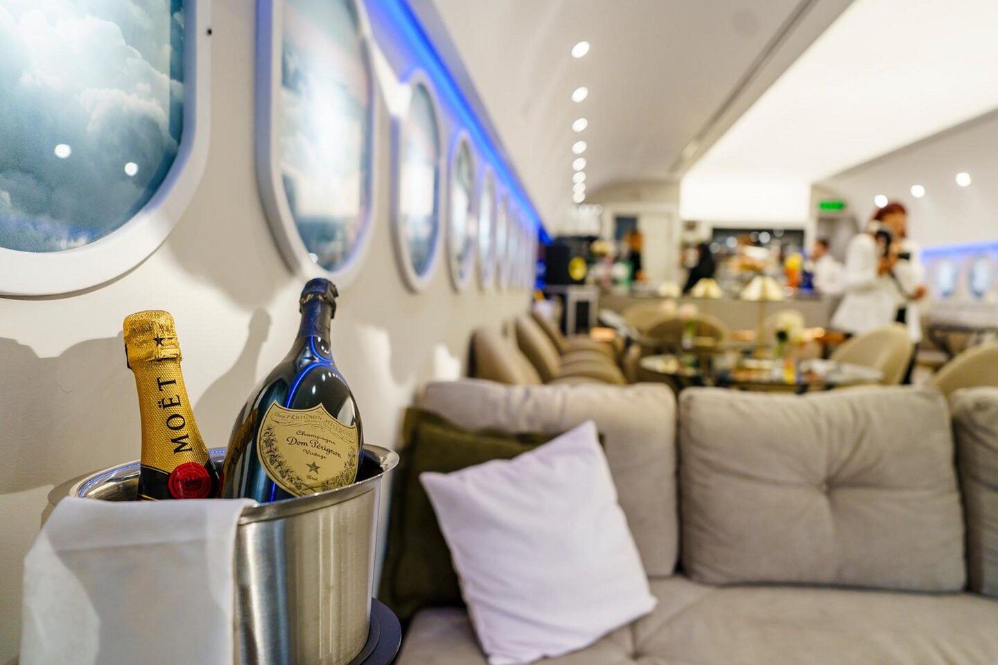 Lounge corner set for service: Moet and Dom Perignon bottles in a steel ice bucket on a linen-dressed stand, deep cream sofa with cushions, backlit portholes and blue cove receding to the bar.