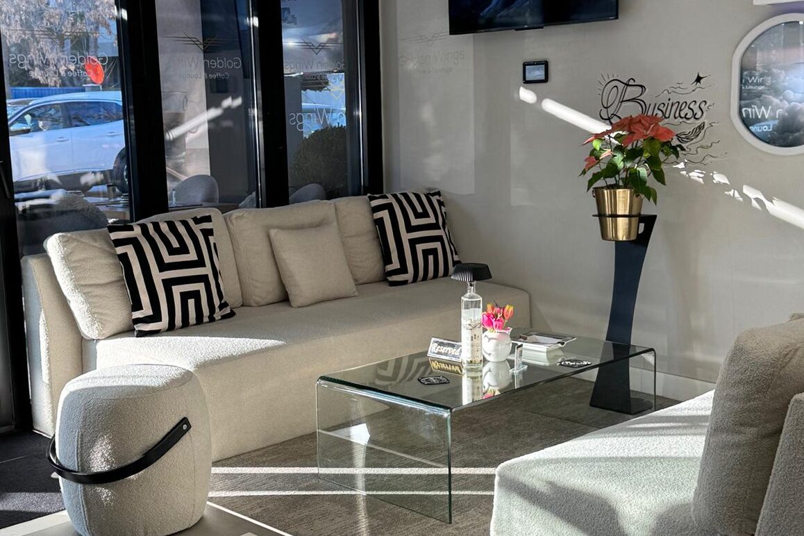 Lounge seating by the glazed end wall: long cream sofa with graphic cushions, glass coffee table, wall-mounted screen under the curved ceiling, a porthole to the right and runway markings on the floor.