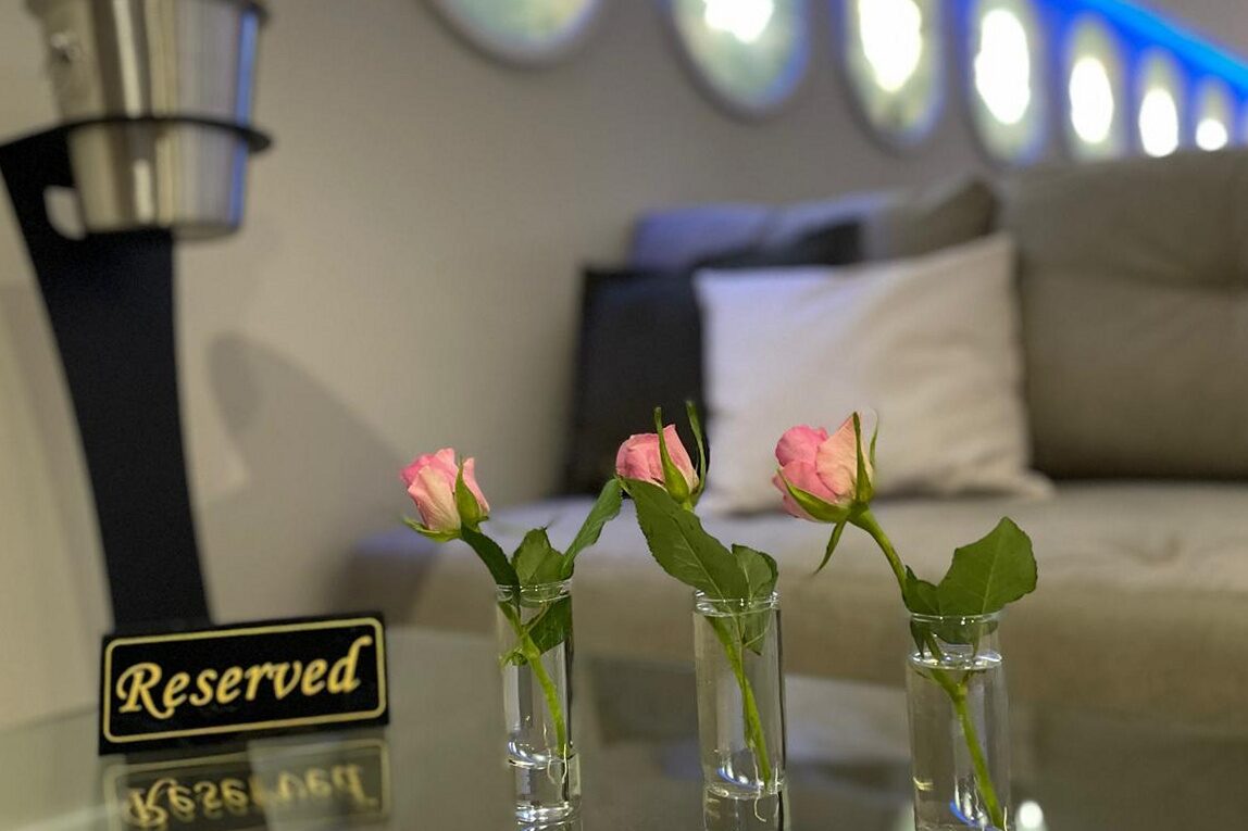 Table detail: a brass Reserved plate and three single roses in glass stem vases on a glass table top, ice bucket stand behind, backlit portholes and blue cove out of focus beyond.
