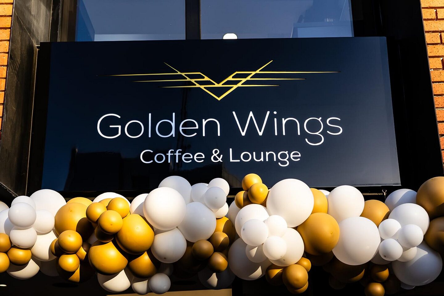 Opening night: the illuminated Golden Wings Coffee and Lounge sign with its gold chevron mark, dressed with a gold and white balloon garland against the brick wall.