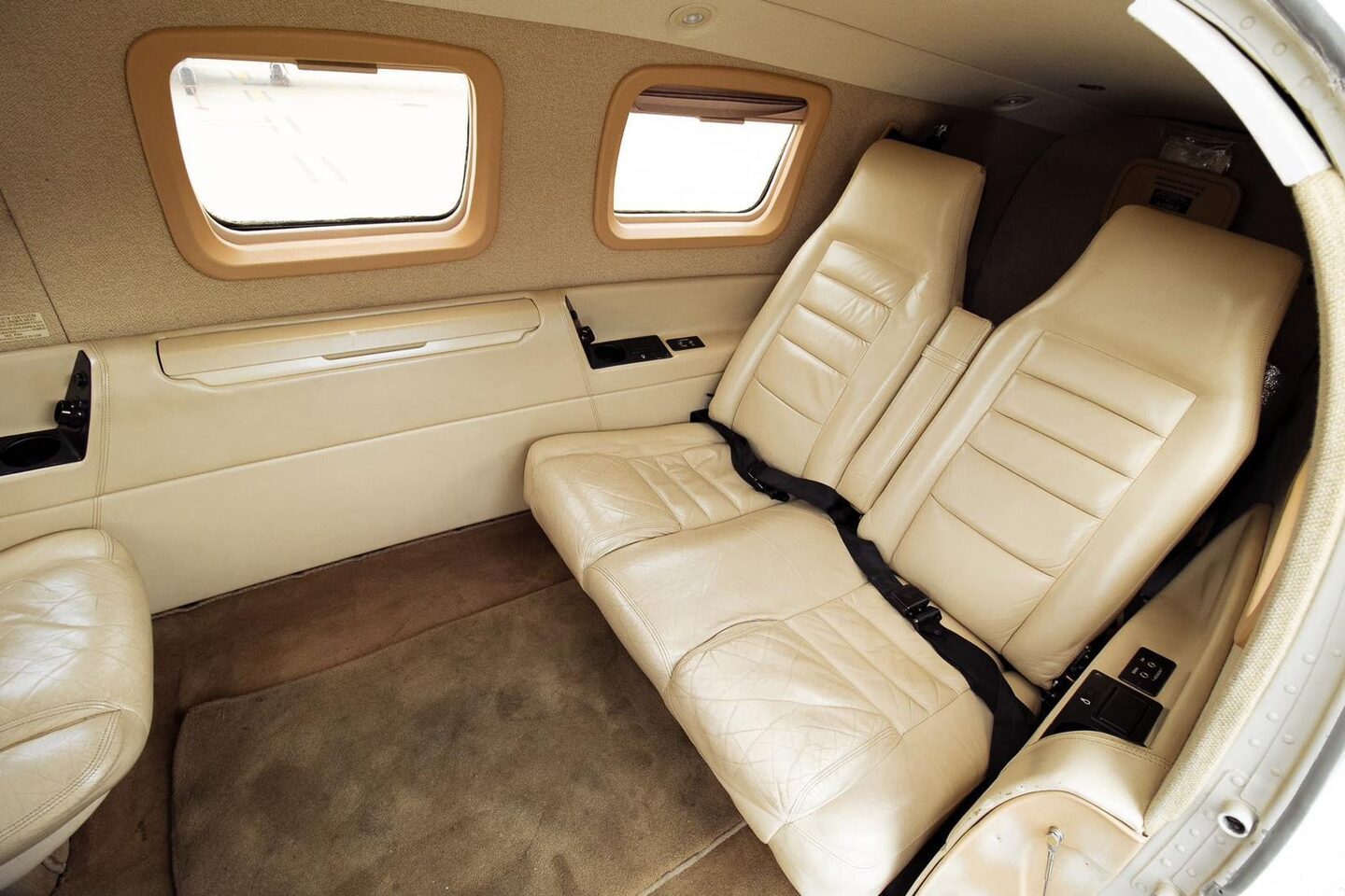 Original Malibu cabin aft section: two-place cream leather bench with headrests below a pair of square cabin windows, tan sidewall trim, worn carpet.