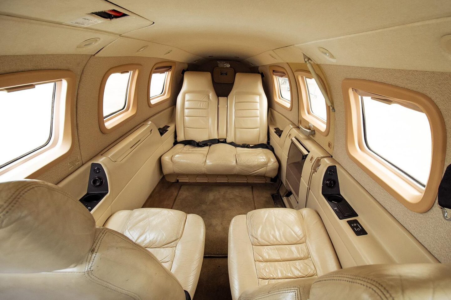 Original Piper PA-46 Malibu cabin looking aft: four yellowed cream leather club seats facing each other across a folding table stub, rear bench under the aft window, tan carpet and matching tan sidewall panels.