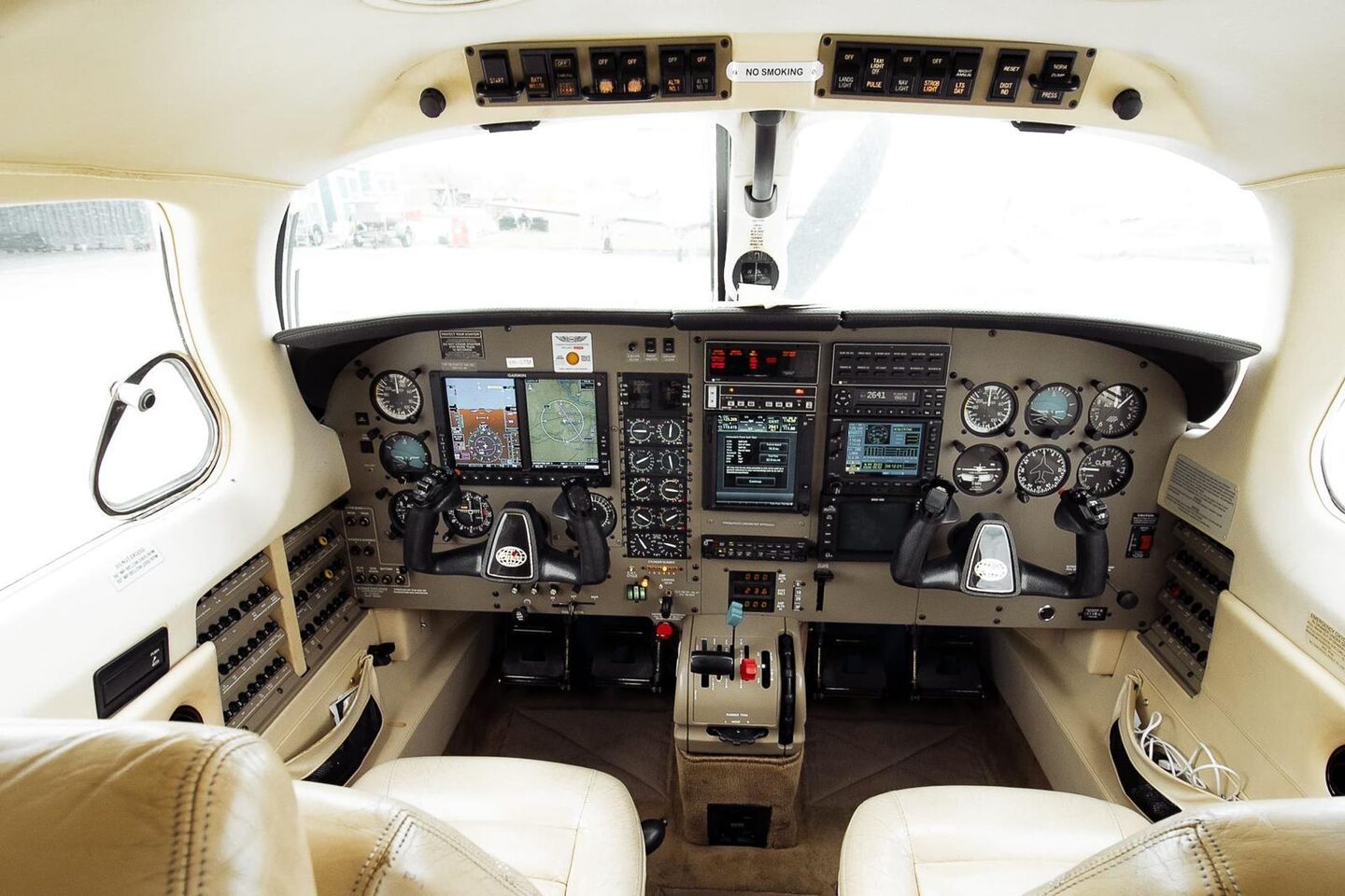 Original Malibu flight deck: twin control yokes, Garmin glass displays flanked by round analogue instruments, cream leather pilot and co-pilot seats, tan overhead switch panel.