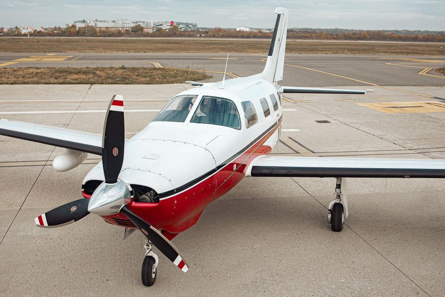 The aircraft on the apron before refit: white Piper PA-46 Malibu with red and black cheatline, three-blade propeller, airfield and treeline behind.