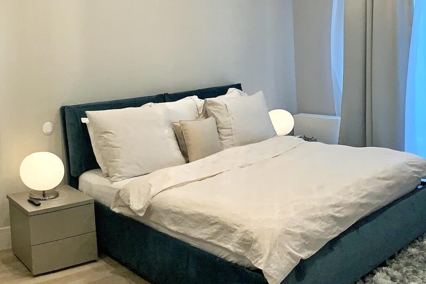 Bedroom: teal velvet upholstered bed with white linen, matching bedside cabinets with globe lamps, floor-length curtains, oak floor and a pale rug.