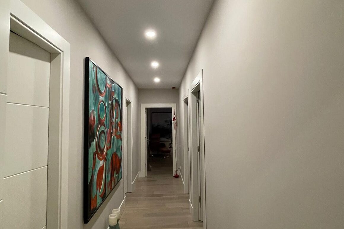 Hallway: white walls and flush doors with a run of recessed downlights along the ceiling, a framed abstract canvas on the left wall, oak plank floor running to the far end.