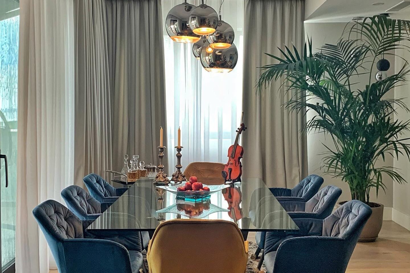 Dining room: glass-topped table with blue velvet chairs and one tan leather head chair on a deep-pile rug, smoked-glass cluster pendant above, floor-length curtains and a palm at the window.
