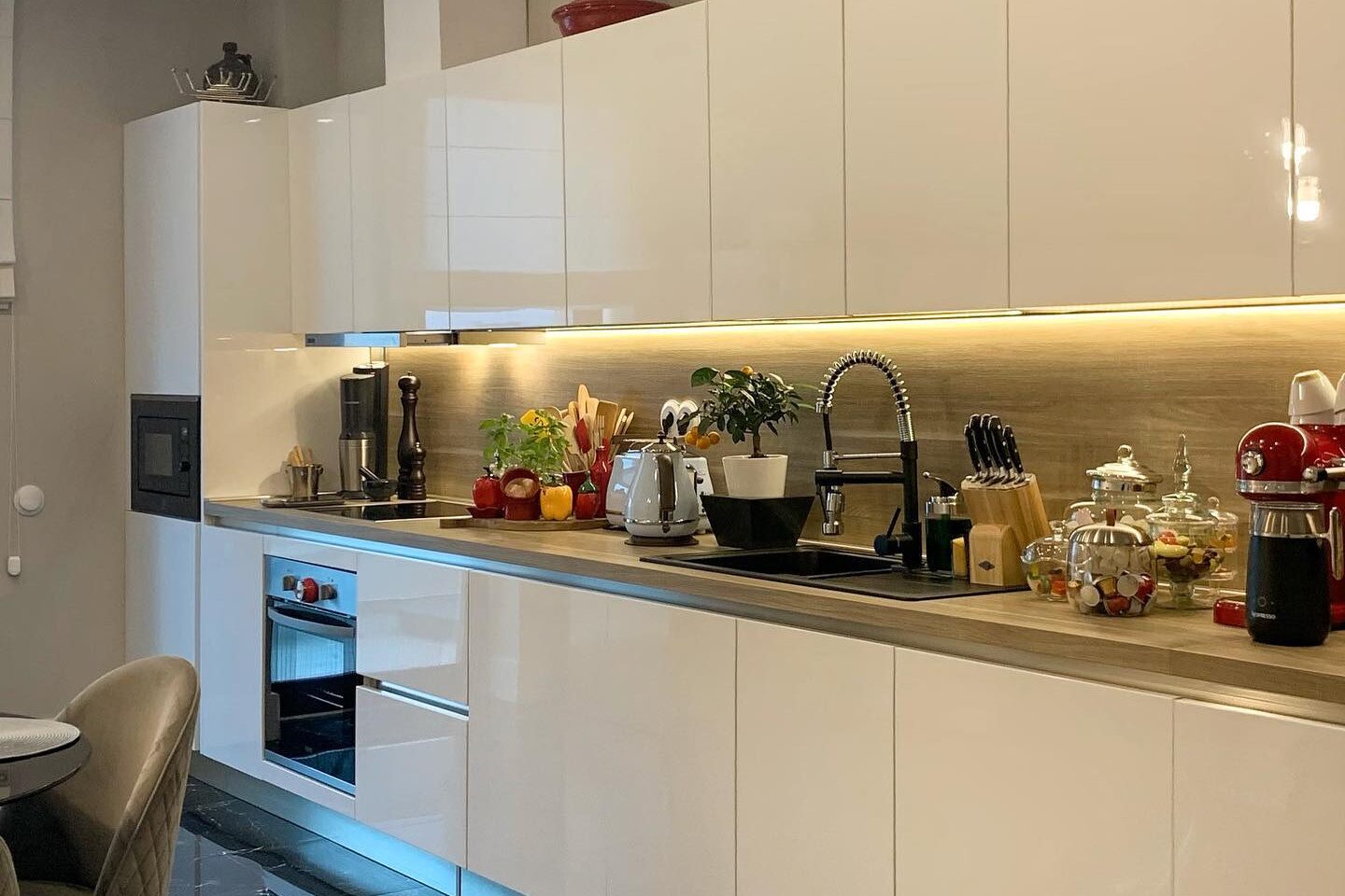The full kitchen run: gloss white wall and base units with an oak worktop and under-cabinet lighting, sink and hob in line, appliances and glass storage jars on the counter, polished black marble floor.