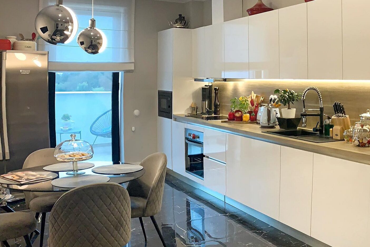 Kitchen and dining: handleless gloss white units with an oak worktop and lit splashback, integrated oven column, polished black marble floor, round dining table with quilted chairs under chrome sphere pendants.