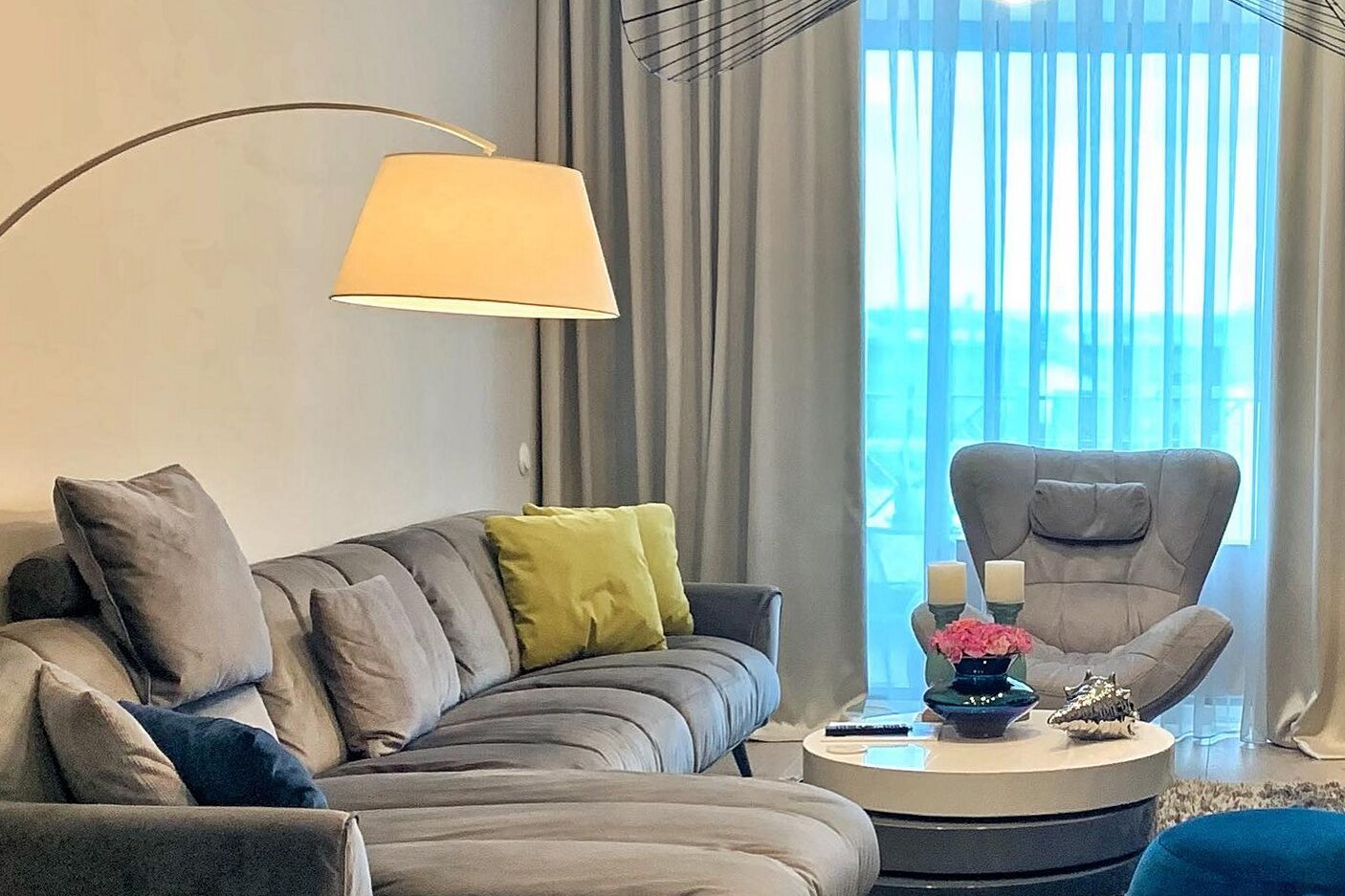 Living room: L-shaped grey sofa with mustard and navy cushions, arc floor lamp over a low round table, black spoke pendant, swivel armchair by the curtained window, oak floor and deep-pile rug.