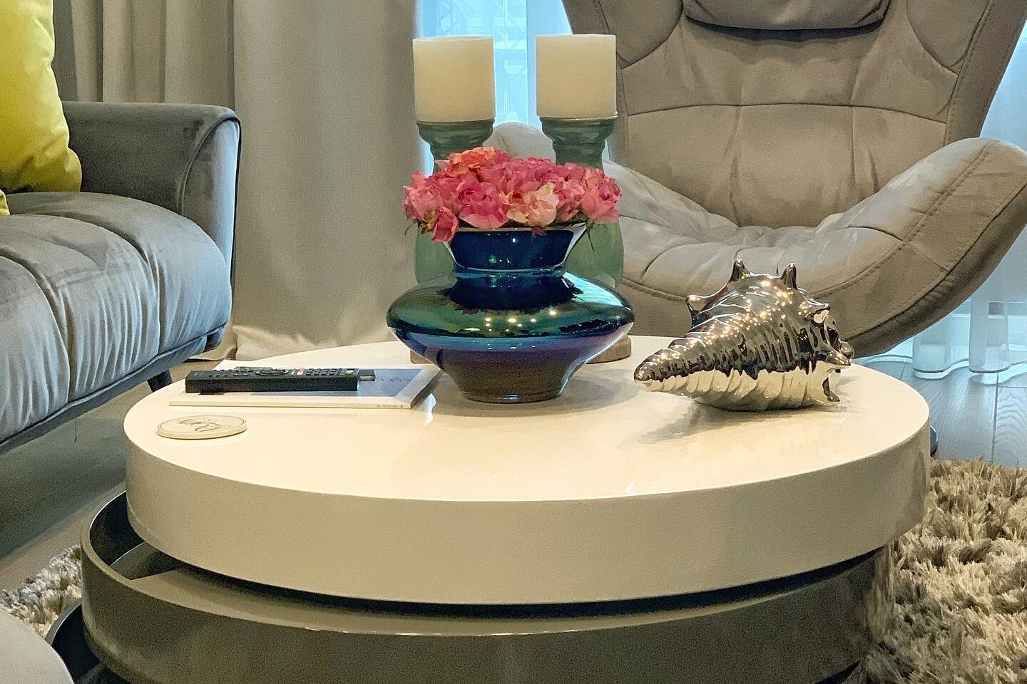 Living room detail: stacked round nesting coffee tables in cream and olive on a shaggy rug, a shell and a blue ceramic bowl of pink blooms on top, grey sofa and high-backed swivel chair behind.