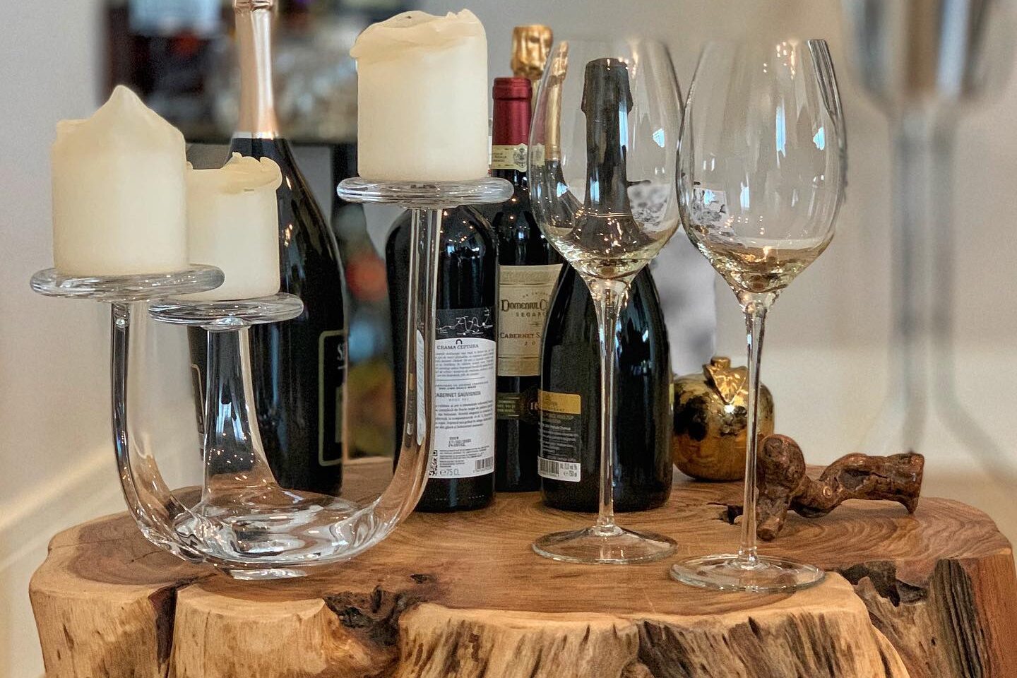 Detail: a solid walnut burl slab table on castors dressed with wine and champagne bottles, crystal glasses and pillar candles, bar shelving out of focus behind.
