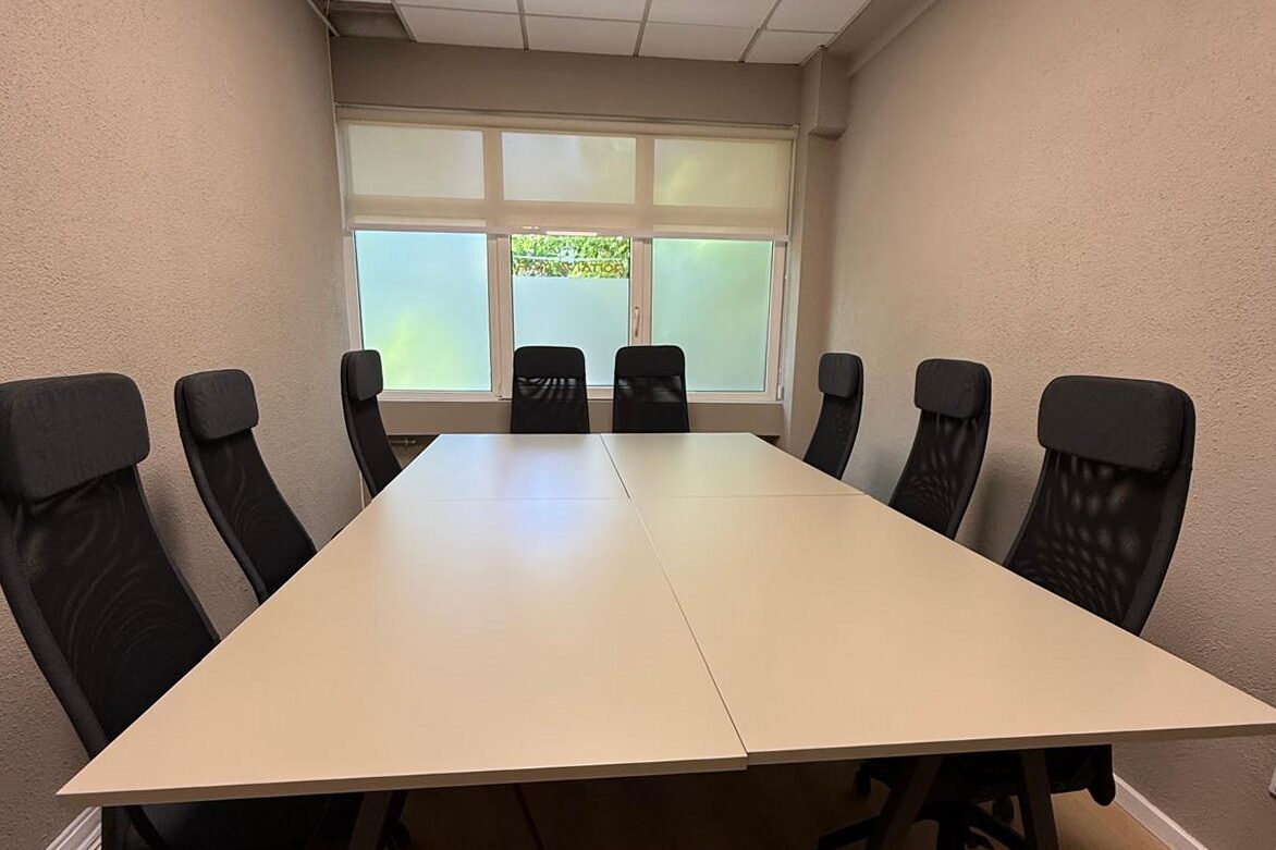 Boardroom: long light-oak meeting table with eight black mesh task chairs, roller blinds across the window wall, oak plank floor.