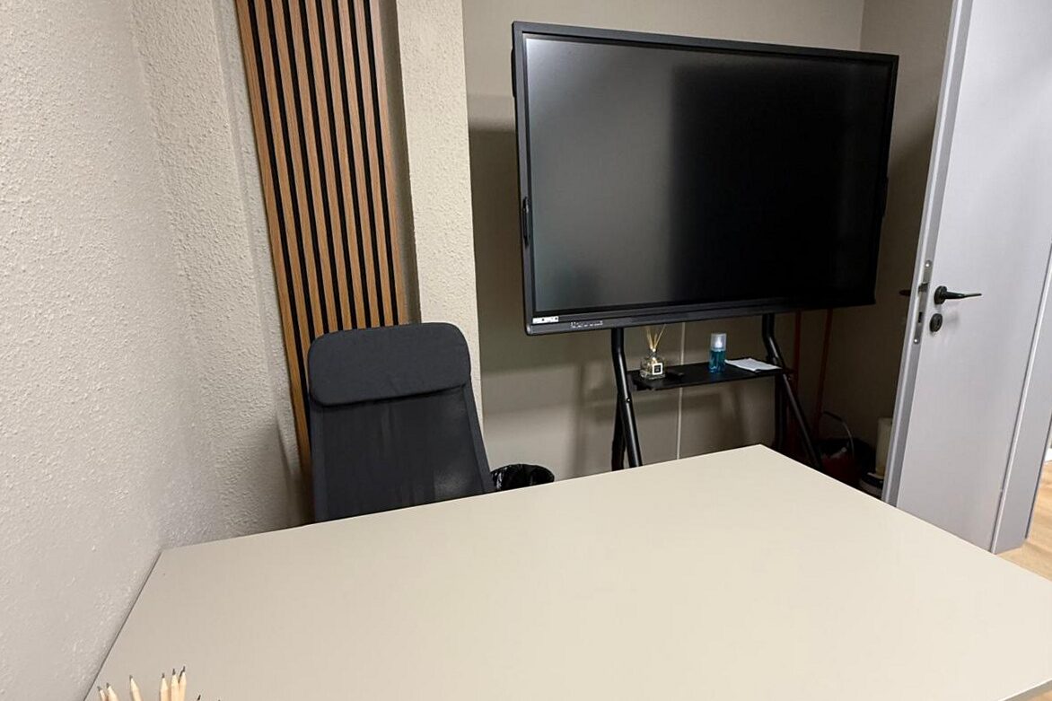 Breakout and presentation corner: white work table on black legs, wall screen on a stand, oak slat panel running full height on the left, oak plank floor.