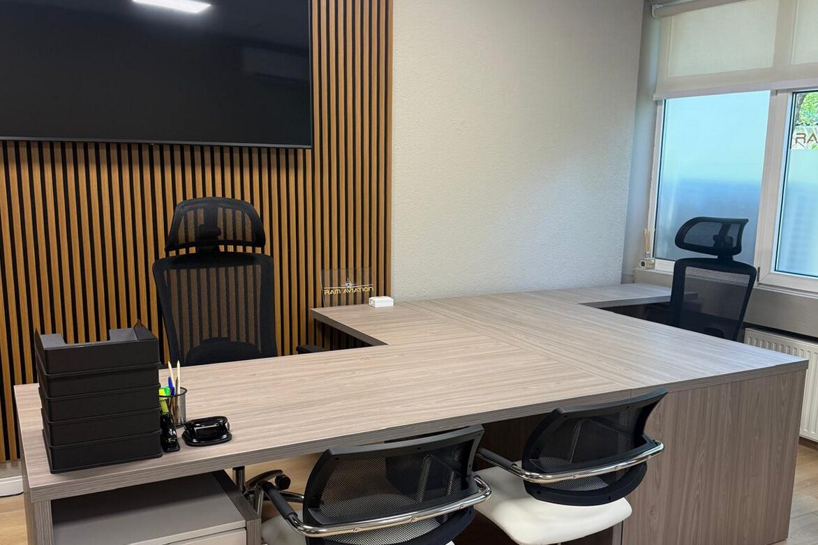 Director's office: L-shaped oak-veneer desk with white cantilever visitor chairs, vertical oak slat feature wall carrying a large wall-mounted screen, oak plank floor, window to the right.