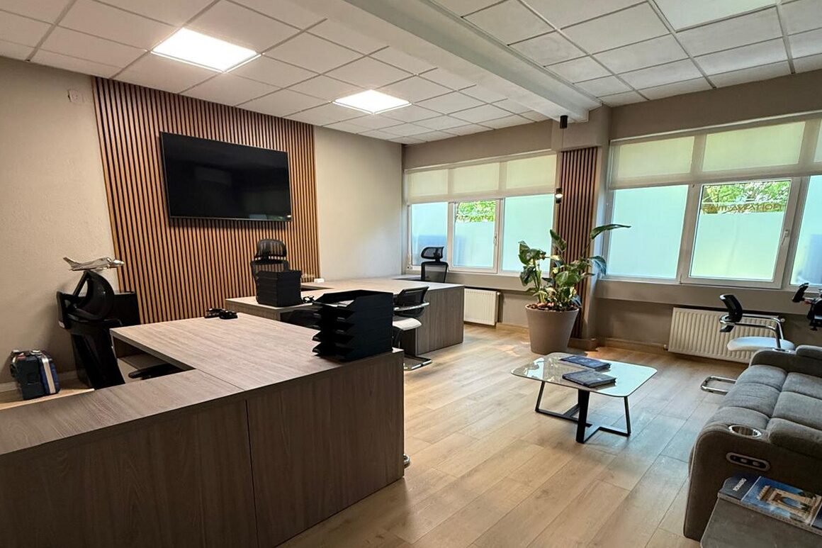 Open-plan office from the entrance: oak-veneer reception counter in the foreground, oak slat panel and screen on the far wall, desks along the window run, soft seating and coffee table to the right.