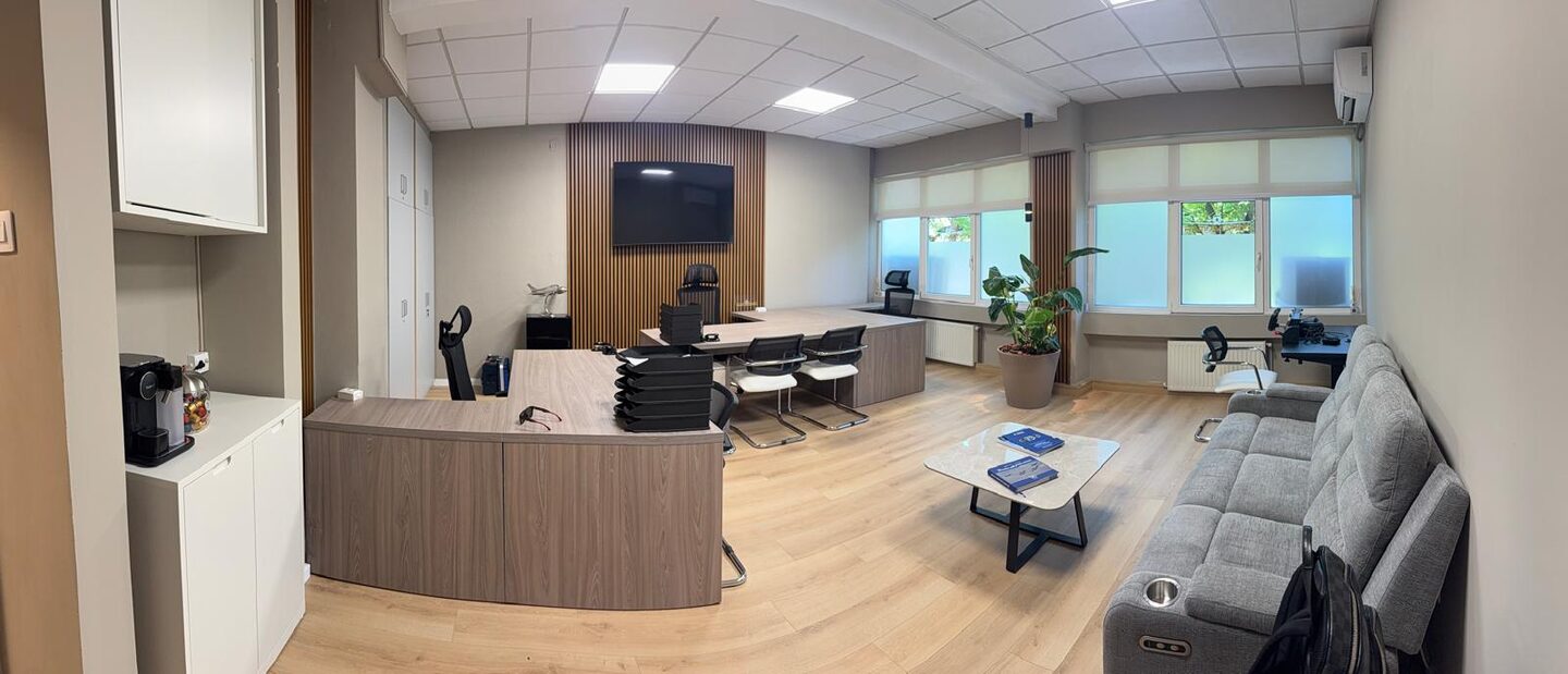 Panorama of the whole office floor: coffee point and reception counter at the left, oak slat wall and screen at the centre, desking along the windows, sofa and lounge seating at the right.